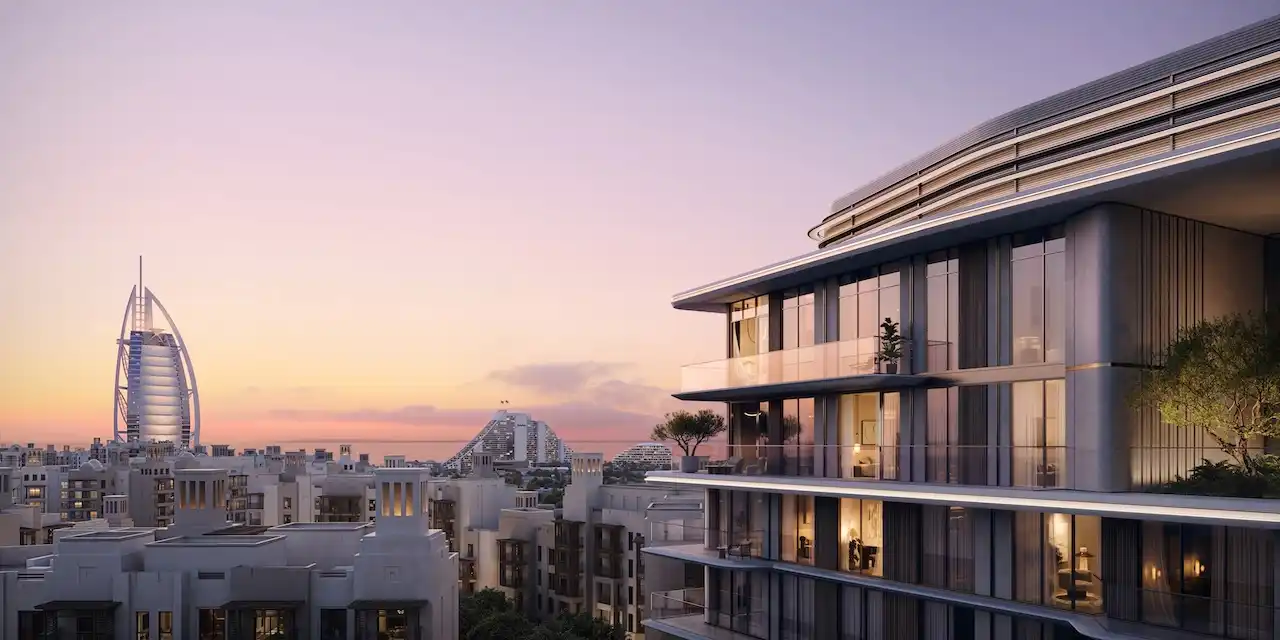 Exterior sunset view of MJL Nourelle in Madinat Jumeirah Living Dubai, showcasing elegant contemporary architecture with illuminated balconies and Burj Al Arab views, set within a low rise community near the Gulf. Presented by Tohid Fetrat.
