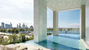 Infinity swimming pool at Atelis at d3 overlooking a serene waterfront and Dubai skyline, featuring marble-clad columns, shaded poolside seating, and panoramic water views in Dubai Design District. Presented by Tohid Fetrat.