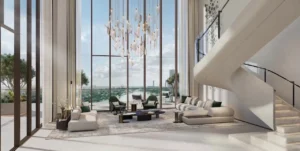 Double-height sky villa living room at Atelis at d3 featuring floor-to-ceiling windows, sculptural staircase, designer chandelier, and panoramic views over Dubai Design District. Presented by Tohid Fetrat.
