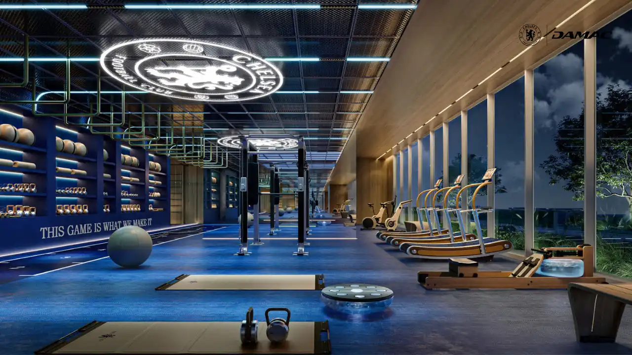 Interior view of the Chelsea Residences athlete training centre featuring a premium gym with professional fitness equipment, branded design elements, and floor to ceiling windows overlooking landscaped surroundings in Dubai, Presented by Tohid Fetrat