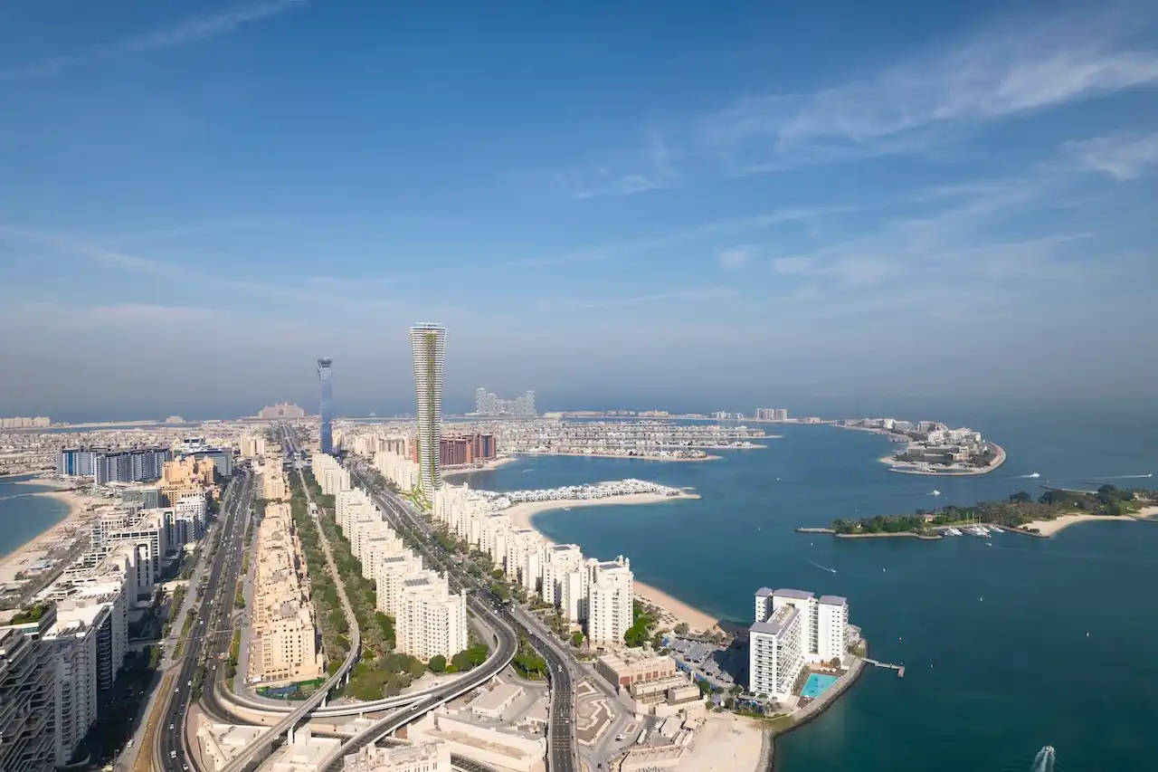 Aerial view of COMO Residences rising along the Palm Jumeirah trunk, showing the tower’s slender vertical form, surrounding beachfront communities, road network, and expansive Gulf waters. Presented by Tohid Fetrat.