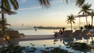 Waterfront infinity pool at COMO Residences featuring a calm reflective pool edge, elegant sun loungers, palm trees, and a serene sunset view overlooking the sea and Dubai skyline from Palm Jumeirah. Presented by Tohid Fetrat.