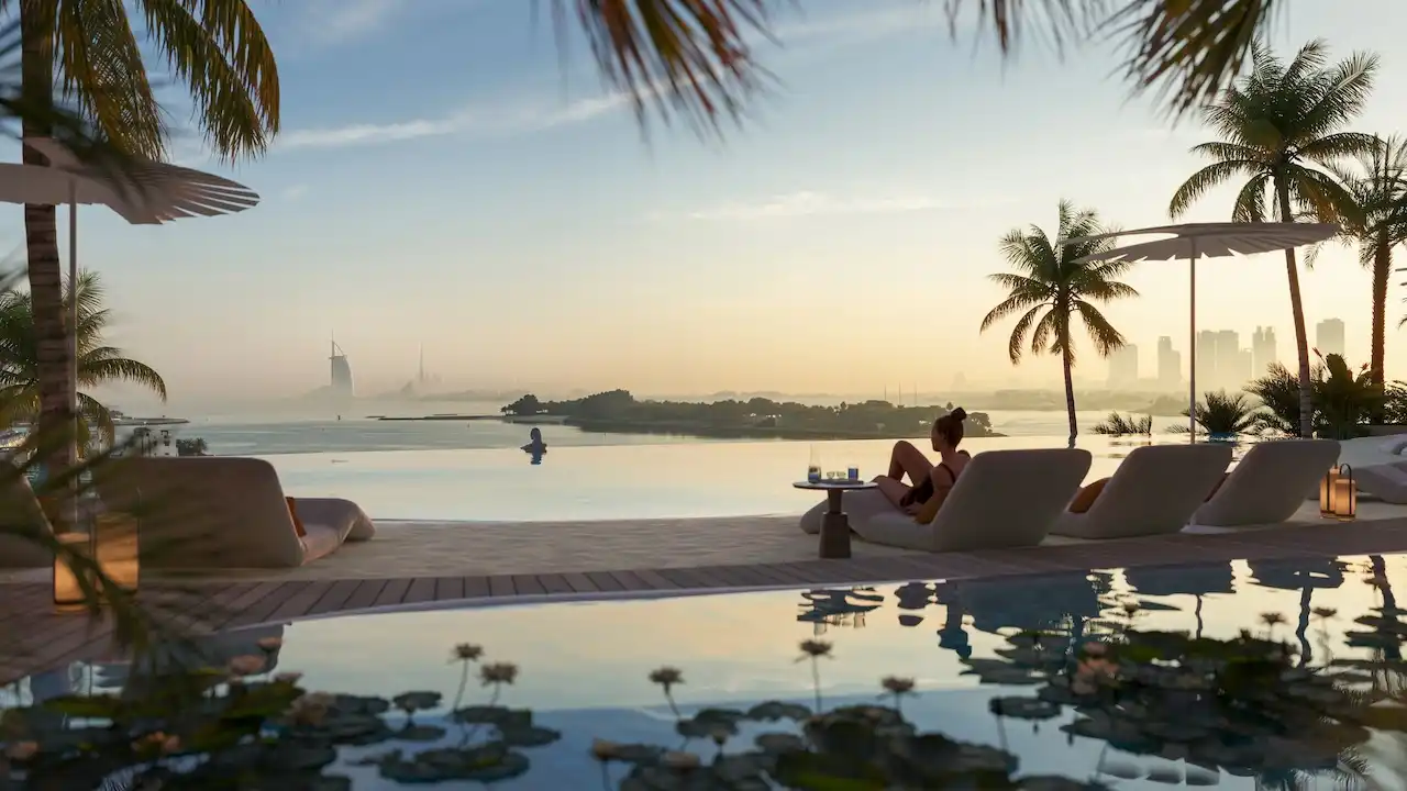 Waterfront infinity pool at COMO Residences featuring a calm reflective pool edge, elegant sun loungers, palm trees, and a serene sunset view overlooking the sea and Dubai skyline from Palm Jumeirah. Presented by Tohid Fetrat.