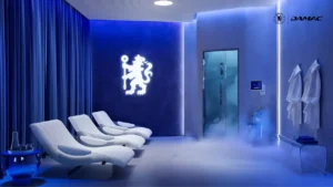 Interior view of the cryotherapy wellness room at DAMAC Chelsea Residences featuring sculpted lounge chairs, ambient blue lighting, branded Chelsea emblem, and a modern recovery setting in Dubai, Presented by Tohid Fetrat