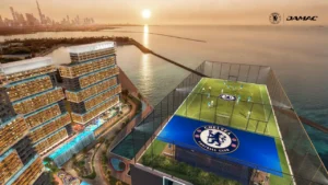 Aerial view of The Stamford Summit rooftop football pitch at DAMAC Chelsea Residences featuring a full size training field with Chelsea branding, elevated netted enclosure, and panoramic waterfront and skyline views at sunset in Dubai, Presented by Tohid Fetrat