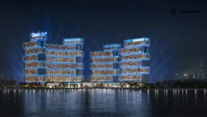 Nighttime waterfront view of DAMAC Chelsea Residences illuminated with the Victory Beacon celebration light show, featuring glowing architectural facades, synchronized beams of light, and reflections across the water in Dubai, Presented by Tohid Fetrat