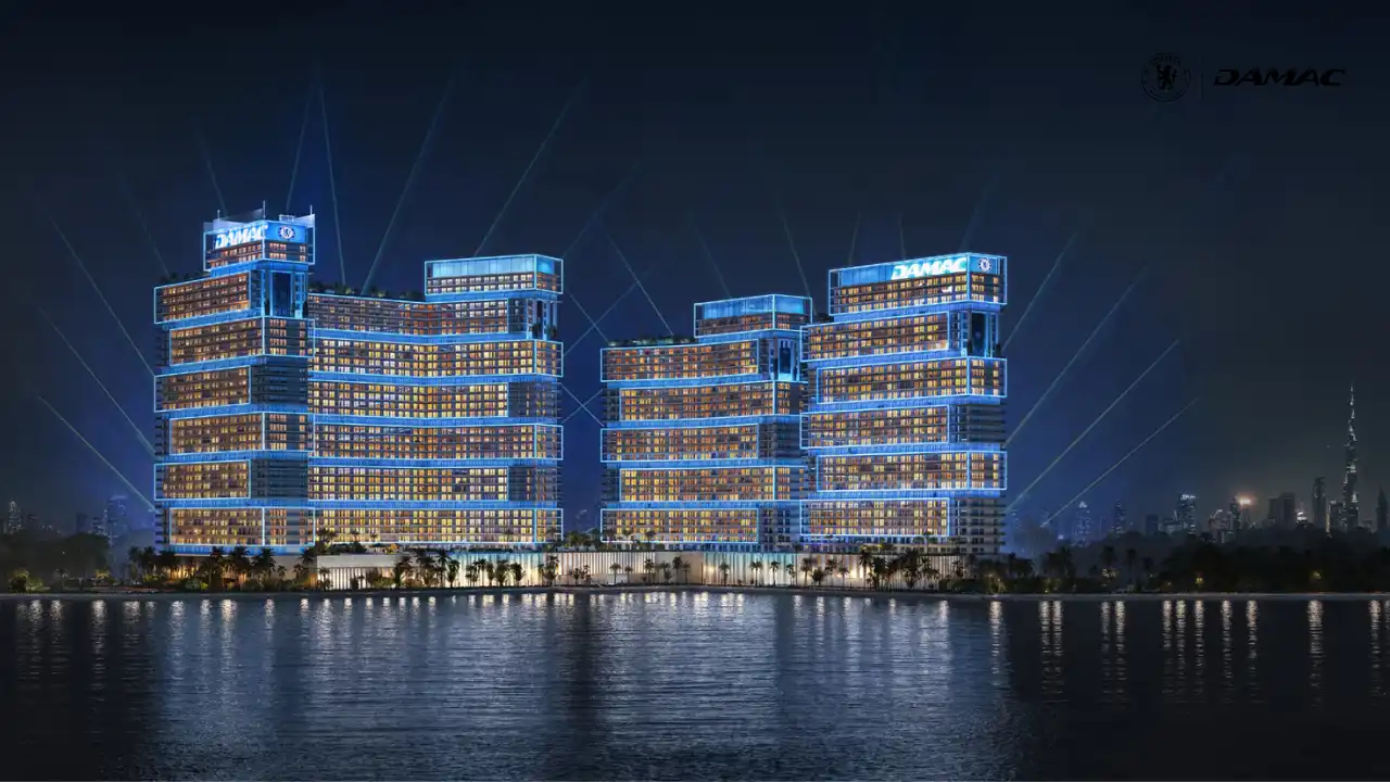 Nighttime waterfront view of DAMAC Chelsea Residences illuminated with the Victory Beacon celebration light show, featuring glowing architectural facades, synchronized beams of light, and reflections across the water in Dubai, Presented by Tohid Fetrat