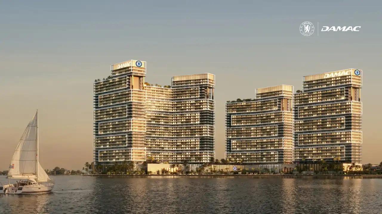 Waterfront exterior view of DAMAC Chelsea Residences featuring contemporary stepped towers, expansive balconies, Chelsea branding, and a calm coastal setting with open water and skyline views in Dubai, Presented by Tohid Fetrat