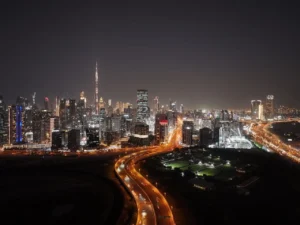 Aerial front night view of Haus of Tenet by IRTH in Business Bay with the Burj Khalifa skyline, illuminated highways, and Dubai’s city lights, presented by Tohid Fetrat