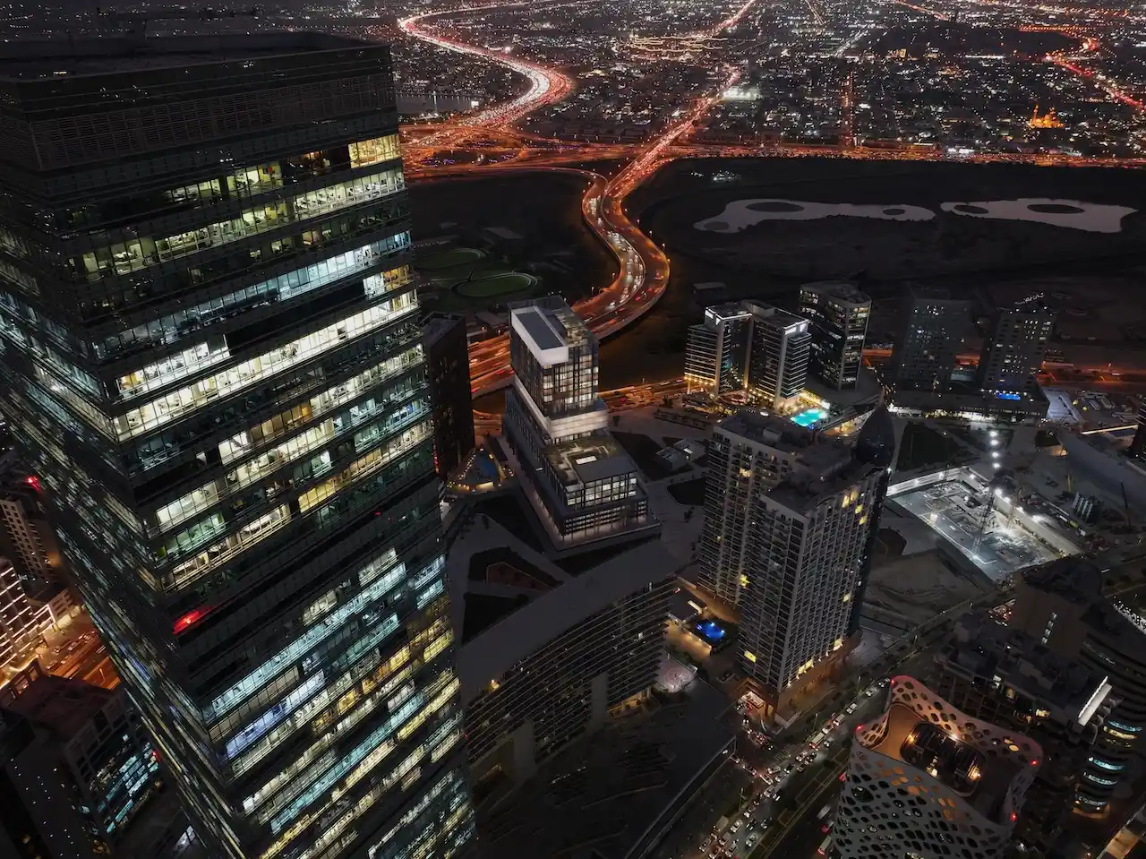 Aerial night view of Haus of Tenet in Dubai with illuminated towers, surrounding skyline, and flowing highway lights across Business Bay, presented by Tohid Fetrat