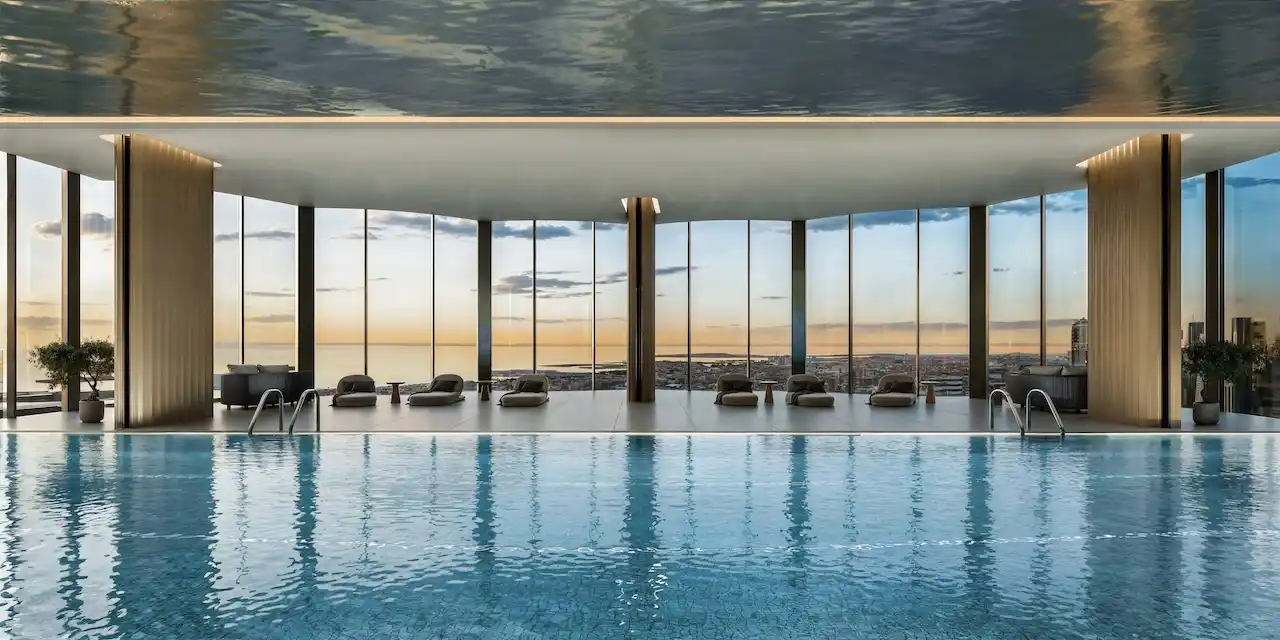 Indoor luxury swimming pool at Lumena Alta featuring floor to ceiling glass walls panoramic city views refined lounge seating and a serene wellness-focused design in Dubai. Presented by Tohid Fetrat.