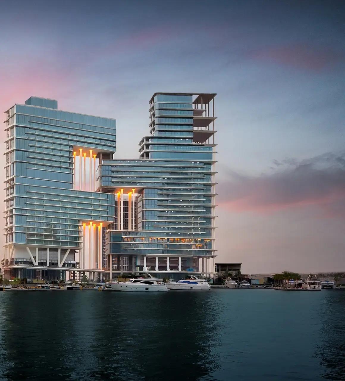 The Lana Residences Dorchester Collection by Omniyat waterfront towers illuminated in the evening with marina and yachts in Dubai - Presented by Tohid Fetrat